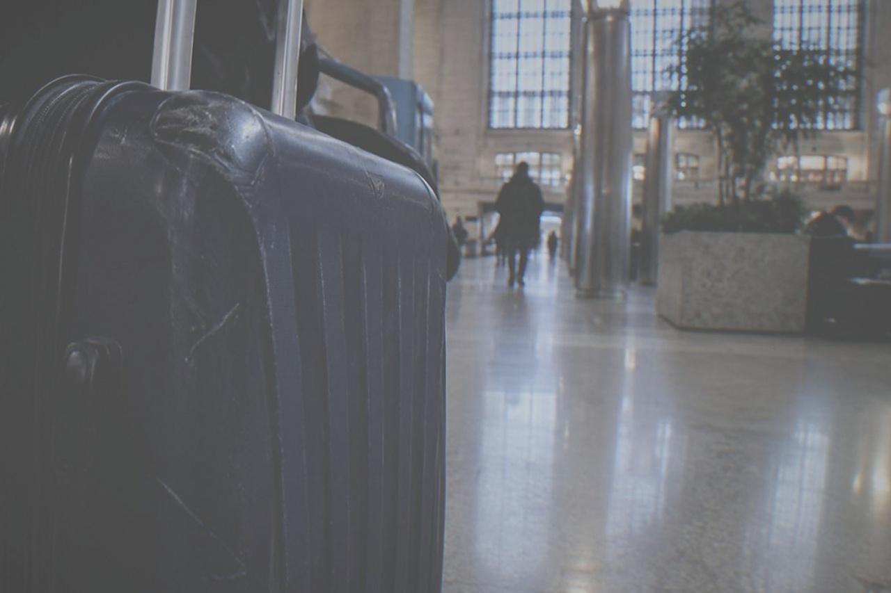 A train hall interior with a wide departure board and travellers walking with wheeled suitcases across a polished stone floor