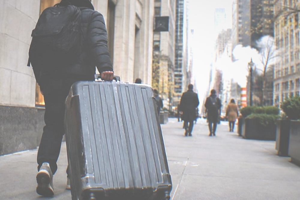 A traveller rolling a large suitcase along a wide New York sidewalk away from a storage shop