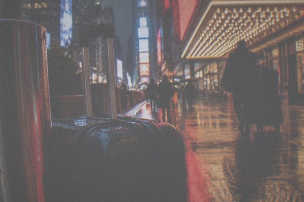 Busy sidewalk in a theatre district with a person pulling a wheeled suitcase past a marquee